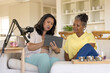 © wavebreak3 - Mature and senior African American women sitting on sofa, showing tablet and prepping microphone