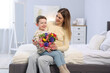 © New Africa - Little boy greeting his mom with flowers on bed at home