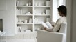 © Nunung - A woman in a cozy sweater sits in a white armchair reading a book in a minimalist living room with a large white bookshelf
