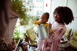© Marko Geber - Mother helping toddler water plants on sunny balcony