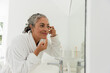 © Wavebreak Media - Senior African American woman leaning at bathroom sink mirror, applying cream near eye in bathrobe