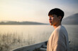 © saltdium - Close portrait of a young Asian man standing by a peaceful lake at sunrise, looking into the distance with warm sunlight on his face, expressing quiet confidence and reflective mood