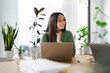 © Studio Marmellata - A young woman smiles while working on her laptop in a bright, modern office space with plants.