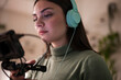 © Studio Marmellata - A young woman wearing headphones is focused on a microphone, preparing to record audio or a podcast.