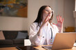 © Studio Marmellata - A young female doctor in a white coat and stethoscope gestures while speaking, with a laptop open on the desk in front of her.