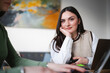© Studio Marmellata - A young woman with dark hair rests her chin on her hand while looking at the camera, holding a phone. Another person is visible in the foreground, working on a laptop.