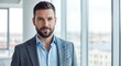 © MopoWe - Bearded professional man wearing a grey textured blazer and light blue shirt stands in a bright modern corporate office with large glass windows