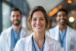© EricMiguel - Smiling female doctor in white coat and blue scrubs with stethoscope in a bright hospital corridor with two blurred colleagues and warm bokeh lights