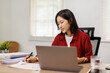 © David - Young Asian woman working from home, focused on laptop while analyzing financial spreadsheets and paperwork at a wooden desk, taking notes and reviewing documents