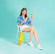 © sitthiphong - Cheerful young Asian woman in a floral shirt sits in a deckchair holding a Thai passport and a credit card and a smartphone on blue background, Concept Songkran Festival in Thailand