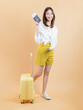 © sitthiphong - Happy young Asian woman broken arm wrapped holding passport with yellow suitcase smiling on orange background.