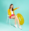 © sitthiphong - Cheerful young Asian woman enjoys her summer vacation, sitting in a deckchair with a yellow rubber ring and pointing forward with a surprised expression against a vibrant blue background