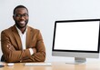 © tutun - Smiling African American businessman in a brown suit sitting at a desk with a computer.