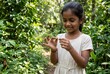 © Chaery - Smiling Indian girl holding small plant in lush garden, environmental awareness and nature concept