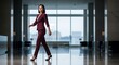 © Lucianastudio - Confident businesswoman in a stylish maroon suit walking purposefully through a modern office lobby with large windows and city views
