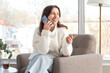 © Pixel-Shot - Young woman talking by mobile phone in armchair near large window at home