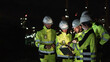 © AU USAnakul - Five Construction Workers Examine Construction Site with Laptop, Reviewing Plans in Dimly Lit Industrial Area, Wearing Hard Hats and Yellow Vests, Conducting Inspection Work at Nighttime.