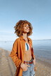 © SHOTPRIME STUDIO - Young woman with curly hair standing on the beach smiling, wearing casual outfit with striped top and orange shirt, enjoying sunny day and fresh sea breeze outdoors.