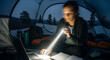 © MstNelufar - Woman using laptop inside tent at night, focused on work with a flashlight beam illuminating the keyboard, enjoying remote work in nature