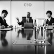 © Liza - Glass of water on a corporate meeting table in an office with blurred business people, CEO sign, and a laptop