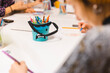 © Francis - Close-up of a creative blue monster-themed pencil holder filled with colorful pencils on a white table during a children's art workshop, with blurry kids drawing and crafting in the background.