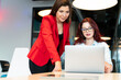 © bulentbaris - A woman in a red blazer assists another woman with glasses at a laptop on a conference table in a modern office. They are coworkers engaged in a work project.