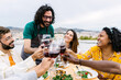 © Xavier Lorenzo - Diverse friends on a rooftop terrace enjoying a joyful summer dinner and bbq, clinking wine glasses, laughing and relaxing with food, city view and sunny urban vibe