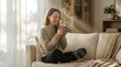 © Yuliia - Young Asian woman in cozy sweater sitting cross-legged on sofa holding warm drink, enjoying peaceful morning moment in bright modern living room.
