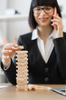 © sofiko14 - A focused businesswoman plays a game of wooden blocks while talking on her mobile phone, symbolizing multitasking and strategic thinking in a professional setting