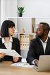 © sofiko14 - A woman and an African American man in business attire collaborate at a desk with documents and a laptop