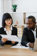 © sofiko14 - A woman and an African American man in business attire collaborate over documents and a laptop in an office setting