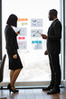 © sofiko14 - A woman and an African American man in suits analyze charts and graphs on a glass wall during a business meeting