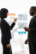 © sofiko14 - A woman in a suit points to charts on a glass wall while a man in a suit listens attentively during a business meeting