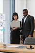 © sofiko14 - A woman and an African American man in business attire collaborate while looking at a laptop screen in a modern office setting