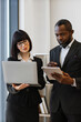 © sofiko14 - A woman and an African American man in business attire collaborate using a laptop and tablet in an office setting