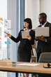 © sofiko14 - A woman in glasses points at charts on a glass wall while a man holds a laptop during a business meeting