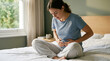 © Moonroad - Young woman sitting on bed feeling intense stomach pain holding abdomen suffering from severe cramps indigestion or menstrual ache expressing discomfort and distress in bright cozy bedroom