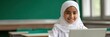 © Iryna - Female asian child in white hijab using laptop in a bright classroom smiling at camera, student learning with technology, education and concentration in school environment