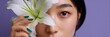 © Iryna - Female asian young adult closeup face with white lily flower covering one eye, green manicure and dewy skin makeup on purple background beauty portrait concept