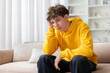 © Anastasiya - Sad bored teen boy in yellow hoodie sitting alone on sofa with hand on cheek, looking down. Lonely depressed young male feeling upset at home