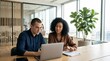 © Phaimanee - Two colleagues, a man in a navy shirt and a woman with curly hair, are focused on a laptop screen at a wooden conference table in a modern, sunlit office with large windows.