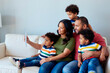 © DragonImages - Wide shot of multiethnic family using tablet while sitting together on sofa, sharing video call or selfie moment. Supporting parenting, family connection, home technology marketing