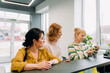© m-art - Three women stand in a bright and inviting reception space, exchanging smiles and ideas. One examines an item closely, while the others listen intently, creating a warm atmosphere.