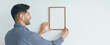 © Prostock-studio - A man stands next to a wall, holding a blank frame with both hands. He is smiling as he prepares to hang the frame for decoration. The setting indicates a home improvement project during the daytime.
