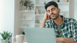 © Prostock-studio - A young man sits at a desk with his hand on his face, appearing tired and distracted. He works on a laptop surrounded by a few items in a well-lit room at home.