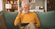 © Stockphotodirectors - An elderly man enjoys his time on the couch. He wears headphones and talks on the phone while holding a tablet. The living room has a cozy setup with soft furniture and warm lighting.