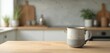 © miss irine - A ceramic mug sits on a wooden kitchen table. The blurred background shows countertops, plants and utensils. This cozy scene invites a warm beverage moment.