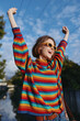 © SHOTPRIME STUDIO - Young woman in a rainbow sweater and yellow sunglasses, smiling with arms raised outdoors by a canal; casual outfit, short hair, carefree travel and lifestyle moment under sunny blue sky.