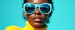 © Viktor - Black woman wears big light blue sunglasses and yellow turtleneck. She poses against bright teal background with sunlight casting shadows. Her look is stylish confident and summery.
