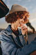 © SHOTPRIME STUDIO - Curly woman wearing beige beanie and denim jacket smiles outdoors during sunset, enjoying a candid moment with soft natural light and blurred background, casual lifestyle vibe